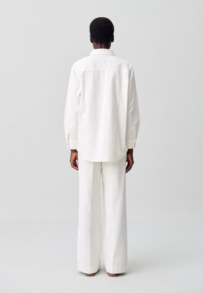 Camicia bianca in cotone con colletto, maniche lunghe e dettaglio sullo yoke posteriore, abbinata a pantaloni bianchi a gamba larga, mostrata da dietro.