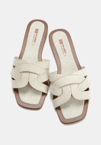 Sandales à enfiler en tissu crème avec un design entrecroisé, semelle plate marron et bout carré. Dessus texturé avec des détails de couture visibles.