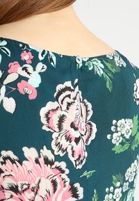 Tissu florale vert avec de grandes pivoines roses et blanches, doté d'une fermeture à bouton dans le dos et d'un détail de couture contrastant.