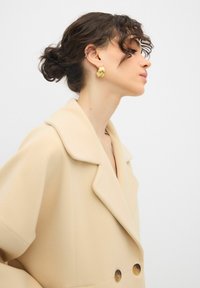 Cappotto beige doppiopetto con grandi revers e bottoni marroni lucidi. Il modello indossa orecchini a cerchio d'oro, mostrando un profilo laterale.