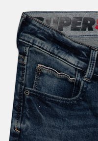 Ciemnoniebieskie dżinsy z lekko wyblakłą teksturą, z kieszenią z przodu i detalami w postaci szwów. Pas ma wyświetloną nazwę marki.