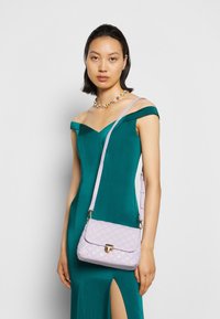 Borsa a tracolla rifinita in lilla con pattina curva, chiusura dorata e tracolla regolabile. Il modello indossa un vestito verde con scollatura off-shoulder, che mette in mostra uno spacco laterale.