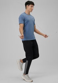 Jeune homme sautant à la corde, portant un t-shirt bleu, un pantalon de sport noir avec une bande grise, et des baskets blanches sur un fond gris.