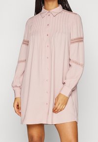 Helles Pink, langärmliges Hemdkleid mit Knopfleiste, das vertikale Falten auf der Vorderseite und Spitzendetails an den Ärmeln aufweist. Weicher, leichter Stoff.