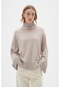 Světlý béžový oversized rolák s ribbenými límcem a manžetami, měkkou texturou a spadlými ramenními švy, doplněný krémovými kalhotami.