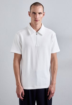 White short-sleeve polo shirt with a ribbed texture, featuring a zippered collar. Worn with navy trousers, model stands against a neutral background.