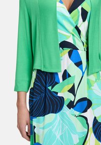 Cardigan léger vert avec des manches trois-quarts porté sur une robe à imprimé floral présentant des motifs bleus, verts et noirs sur fond blanc.