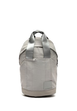 Mochila gris claro con dos asas, bolsillo frontal con cremallera y parche rectangular en la parte inferior derecha, mostrada en posición vertical sobre fondo blanco.