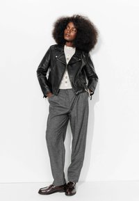 Veste de moto en cuir noir sur un pull léger avec des embellissements, associée à un pantalon à carreaux gris ample et des chaussures noires brillantes.