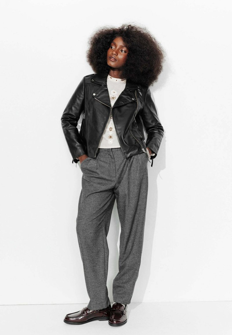 Veste de moto en cuir noir sur un pull léger avec des embellissements, associée à un pantalon à carreaux gris ample et des chaussures noires brillantes.