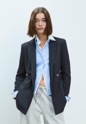 Blazer marine à deux boutons, porté sur une chemise rayée bleu clair, associé à un pantalon de survêtement gris. Tissu lisse, design structuré.