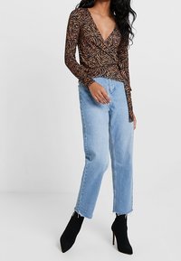 Braun und orange gemusterter langärmeliger Oberteil mit tiefem V-Ausschnitt, kombiniert mit hellblauen hochgeschnittenen Jeans und schwarzen Stiefeletten.