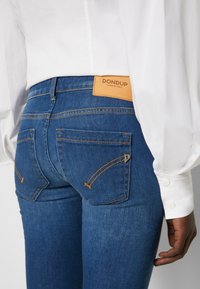 Blå denimjeans med en åtsittande design, orange stickningar och en lädermärke på midjan. Har två bakfickor.