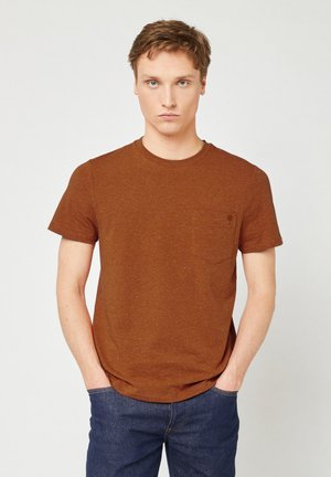 Jonge man met lichtbruin haar, gekleed in een bruine T-shirt met korte mouwen en blauwe jeans, staande met de handen in de zakken tegen een effen achtergrond.