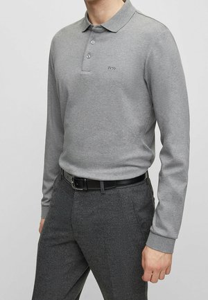 Homme portant un polo à manches longues gris clair avec des boutons, un pantalon gris foncé et une ceinture en cuir noire.
