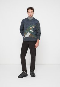 Jersey de punto en azul oscuro con una superficie texturizada y un gráfico de dinosaurio. Combinado con pantalones oscuros y zapatillas negras.