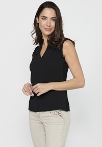 Top senza maniche nero con scollatura a V, caratterizzato da dettagli in pizzo testurizzato sulle spalle. Abbinato a pantaloni beige chiaro decorati con ricami floreali.