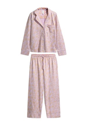 Pitkät tummat hiukset omaava nainen, joka pukeutunut vaaleanpunaiseen pyjamapaitaan, jossa on kultaisia paisley- ja lyhtykuvioita, beige tausta.