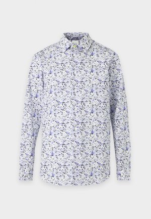 Homme debout portant une chemise blanche à fleurs à manches longues, un pantalon marron foncé et des mocassins jaunes sur un fond uni clair.