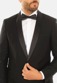 Giacca da smoking nera con rever in raso, abbinata a una camicia bianca e a un papillon nero. Tessuto liscio con una sottile texture.