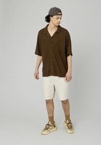 Chemise brune à manches courtes avec un fini texturé ; associée à un short beige clair et à des baskets beiges, avec des chaussettes à motifs.