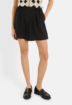 Mujer con pantalones cortos negros texturizados con bolsillos, una camiseta estampada y mocasines negros brillantes, de pie con una mano en el bolsillo.