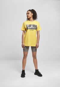 Gul bomullströja med en svartvit grafisk tryck av sammanflätade händer och ordet "be", parat med grå cykelshorts och svarta sneakers.