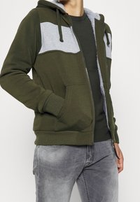 Uomo con felpa oliva e grigia non zipata, foderata in pile, camicia verde oliva e jeans grigi sbiaditi, in piedi con le mani in tasca.