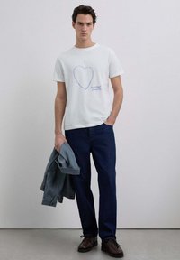 Camiseta de algodón blanca con gráfico de corazón azul y texto "I ♥ SCALPERS company", combinada con vaqueros azul oscuro y una chaqueta azul claro.