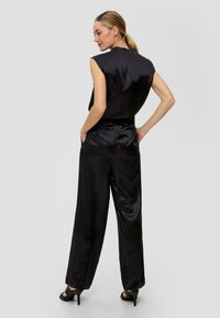 Jumpsuit en satin noir avec des manches courtes, des jambes larges et une fermeture éclair au dos. Texture lisse avec un subtil éclat. Porté avec des sandales à talons hauts noires.