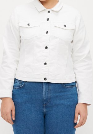 Femme portant une veste blanche boutonnée avec deux poches poitrine et un jean bleu, montrée du cou aux hanches, les mains détendues sur les côtés.