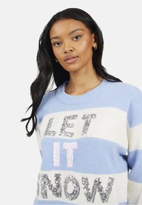 Lichtblauwe en witte gestreepte trui met pailletten tekst "LET IT SNOW" en "IT" in iriserende afwerking. Zachte, textuurstof met ronde hals.