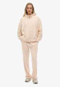 Ensemble de survêtement en velours de couleur pêche comprenant un hoodie zippé avec poches et un pantalon droit assorti, tous deux avec des détails brodés.