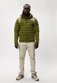 Giacca smanicata verde oliva con zip, pantaloni cargo beige e scarpe da trekking grigie con accenti blu. Persona che porta uno zaino.