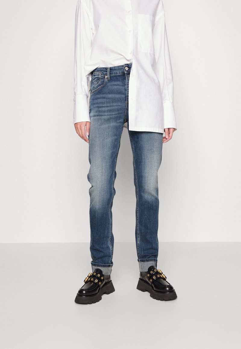 Jeans de mezclilla azul con un ligero desgaste, combinados con una camisa blanca de botones. El calzado es de cuero negro con detalles dorados y suelas gruesas.