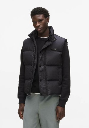 Mann trägt schwarze Steppweste über schwarzem Langarmshirt und hellgrauen Hosen, steht vor einfachem weißem Hintergrund.