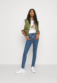 Chaleco acolchado oliva sobre camiseta gráfica blanca, combinado con jeans ajustados azules y zapatillas blancas, contra un fondo claro y liso.