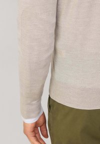 Beżowy sweter z długim rękawem, wełniany, z teksturowanym łatą na lewym ramieniu, noszony na białej koszuli, w zestawie z oliwkowymi spodniami.