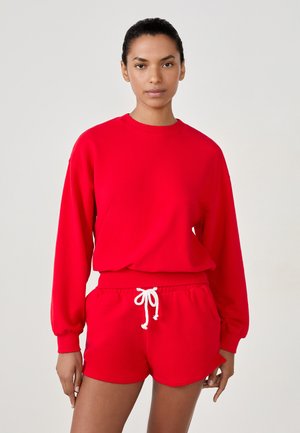 Sweatshirt rouge à manches longues et col ras du cou. Associé à un short rouge avec un cordon blanc. Texture en maille lisse.