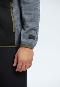 Maglione a maniche lunghe grigio e nero con finitura testurizzata, caratterizzato da un passamano verde oliva e una tasca laterale con cerniera. Logo del brand nero sulla manica.