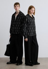 Δύο μοντέλα φορούν όμοια μαύρα πουκάμισα με λευκά κάθετα μοτίβα κειμένου και φαρδιά μαύρα παντελόνια. Συμπεριλαμβάνεται μαύρη τσάντα tote.