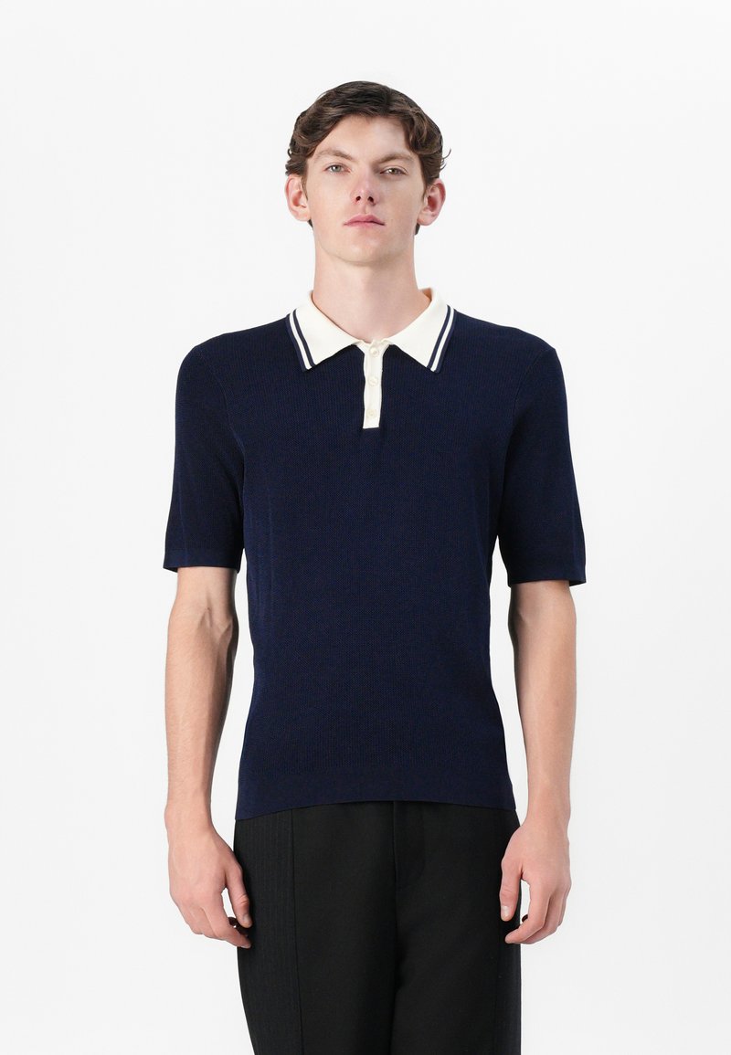 Marineblaues Poloshirt mit kurzen Ärmeln und gerippter Textur, weiß-blau gestreiftem Kragen und drei Knöpfen. Einfaches, tailliertes Design.