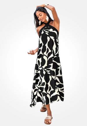 Mujer con vestido halter estampado en blanco y negro, con grandes pendientes y sandalias, posando con una mano en la cabeza y la otra extendida.