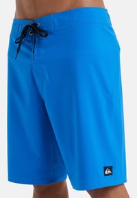 Niebieskie kąpielówki wykonane z lekkiego materiału, z czarnym sznurkiem do regulacji i małą łatką z logo na dolnej lewej nogawce.