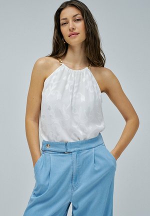Vrouw in een witte mouwloze top met gouden kettingbanden en lichtblauwe high-waisted broek, staand met haar handen in haar zakken.