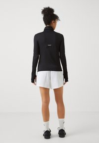 Schwarzes langärmliges Sportoberteil mit hohem Kragen, strukturiertem Stoff und kleinem "TECHFIT"-Logo auf dem Rücken, kombiniert mit weißen Shorts.