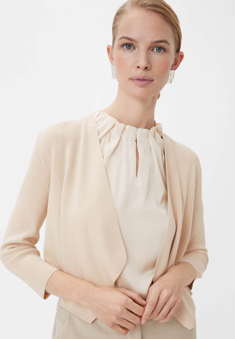 Pfirsichfarbener Cardigan mit weicher Textur, offener Vorderseite und Dreiviertelärmel, kombiniert mit einer cremefarbenen Bluse mit gefälteltem Ausschnitt.
