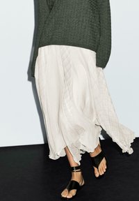 Olivgrüner Strickpullover kombiniert mit einem fließenden, hellen plissierten Rock mit schwachen diagonalen Linien und schwarzen flachen Sandalen.