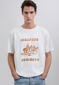 White cotton t-shirt featuring a large, brown graphic print of cowboys on horseback with the text "SCALPERS COWBOYS" below. Classic fit.