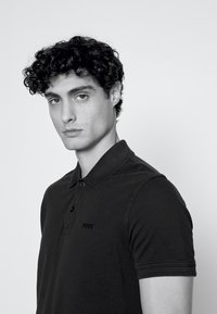 Jaunas vyras garbanotais plaukais dėvi juodą BOSS polo marškinėlį, žiūri į kamerą su neutraliu veidu prieš paprastą foną.